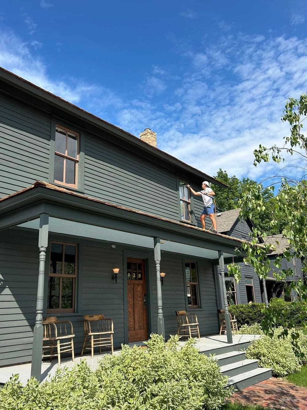 Man painting the roof of a gray house on a sunny day, surrounded by greenery.
